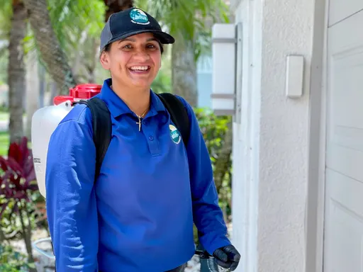 Smiling pest control technician in blue uniform and cap carrying a backpack sprayer at a home entryway.